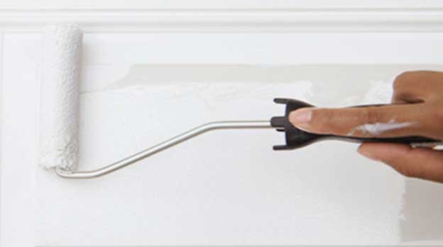 Hand guides a thin roller with white paint across a wall beneath the trim.